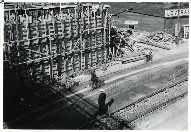 Schwarz-weiß-Foto einer Baustelle mit einem teilweise errichteten Gebäude, Menschen auf Fahrrädern und zu Fuß auf der Straße, Bahnschienen, Strommasten, Holzplanken und andere Baumaterialien, mit Text unten.