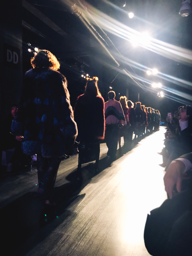 Eine Gruppe von Menschen läuft über einen Laufsteg bei einer Modenschau, beleuchtet von hellen Scheinwerfern, mit Zuschauern auf beiden Seiten.