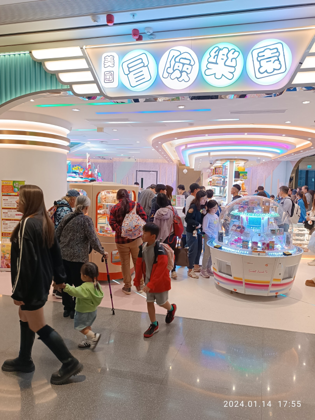 Menschen, die durch ein Einkaufszentrum in Hongkong gehen, tragen einige Taschen und einer hält einen Stock, mit einem Automaten, einer Texttafel, einer Säule und Deckenleuchten im Hintergrund.