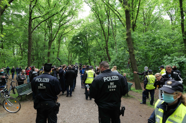 Eine Gruppe von Polizisten steht vor einer Menschenmenge, einige tragen Mützen und Masken, mit Fahrrädern und einer Bank im Vordergrund und Bäumen und Himmel im Hintergrund während einer Anti-Terror-Demonstration in Berlin.