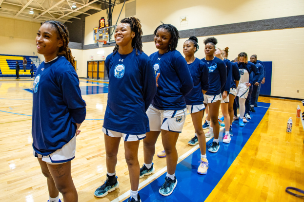 Eine Gruppe junger Frauen in blauen T-Shirts steht lachend auf einem Basketballfeld, mit Flaschen und Gegenständen rechts von ihnen und Zuschauern im Hintergrund.