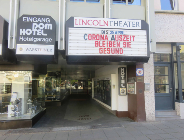 Das Lincoln Theater in Berlin, Deutschland, mit Glasfenstern und -türen und einer Tafel mit Text, sowie einem Inneren, das eine belebte Stadtlandschaft suggeriert.