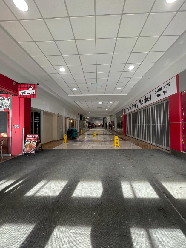 Innenansicht eines Einkaufszentrums mit Deckenbeleuchtung, einem Stuhl und einer Tafel, die "Sydbury Market - Vorher und Nachher" links zeigt, sowie verstreuten Gegenständen rechts und auf dem Boden.