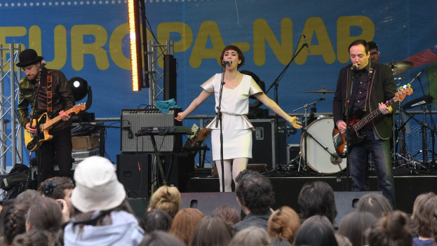 Vier Musiker auf der Bühne: eine Frau in einem weißen Kleid singt in der Mitte, flankiert von zwei Gitarristen, mit einem Publikum unten und Lautsprechern im Hintergrund an der Wand.