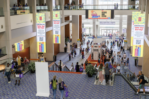 Eine große Gruppe von Menschen, die durch ein großes Gebäude mit Bannern, Topfpflanzen, Rolltreppen, Geländern, Säulen, Texttafeln, Glastüren und anderen Gegenständen geht, wobei einige Taschen tragen, beim Nationalen Buchfestival.