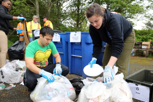Zwei Personen in Handschuhen sammeln Müll auf Tellern in einem Park, umgeben von Müll, mit einem Mülleimer und einer Bank in der Nähe unter Bäumen und einem blauen Himmel.