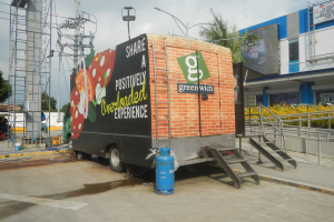 Lastwagen vor einem Gebäude mit einer Tafel, auf der "Teilen Sie eine positiv überladene Erfahrung" steht, mit Stufen, einem Zylinder auf dem Boden, Geländern, Strommasten mit Drähten, Laternen, Bäumen und einem bewölkten Himmel.