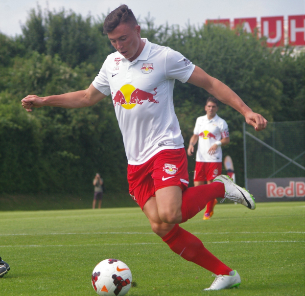 Ein Profifußballer kickt einen Ball auf einem grünen Feld mit "RB Leipzig" im Hintergrund unter einem klaren blauen Himmel.