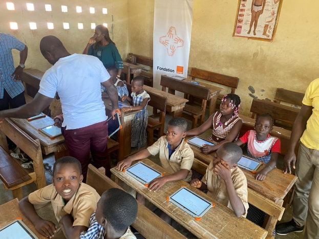 Kinder an Tischen sitzend, die Tablets verwenden, mit Erwachsenen in der Nähe, in einem Klassenzimmer mit einem Banner und einem Plakat an der Wand.