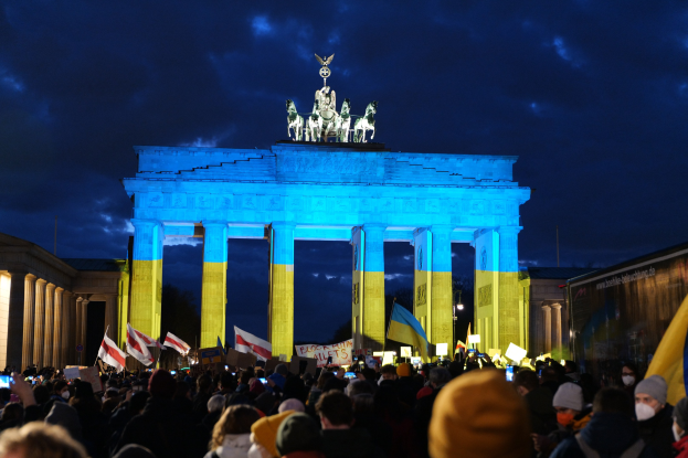 Eine Menschenmenge steht vor dem Brandenburger Tor in Berlin und hält Fahnen und Schilder in den Händen, auf denen ein Banner mit Protesttext zu sehen ist.