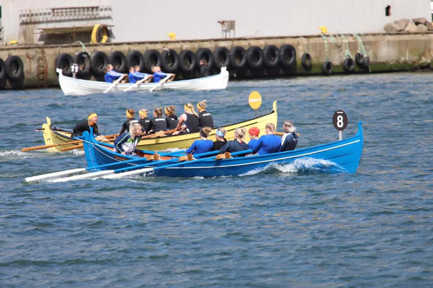 Eine Gruppe von Menschen in einem blauen und gelben Boot auf dem Wasser, die Paddel halten, mit einer Wand mit Reifen und einem Gebäude im Hintergrund, was eine Regatta vermuten lässt.