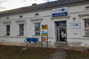 Weißer Bau mit einer Tafel, die "Anipol Post Office" anzeigt, mit Fenstern, einer Tür, einer Tafel mit Text, Pfählen, einem Weg, Gras, trockenen Blättern, einem Baum und einem bewölkten Himmel.