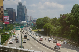 Eine vielbefahrene Autobahn mit zahlreichen Fahrzeugen, gesäumt von hohen Geb├Ąuden, ├ťberlandleitungen, ├Überlandstra├čenbeleuchtung, Werbetafeln und B├Ąumen, unter einem bew├Âlktem Himmel.