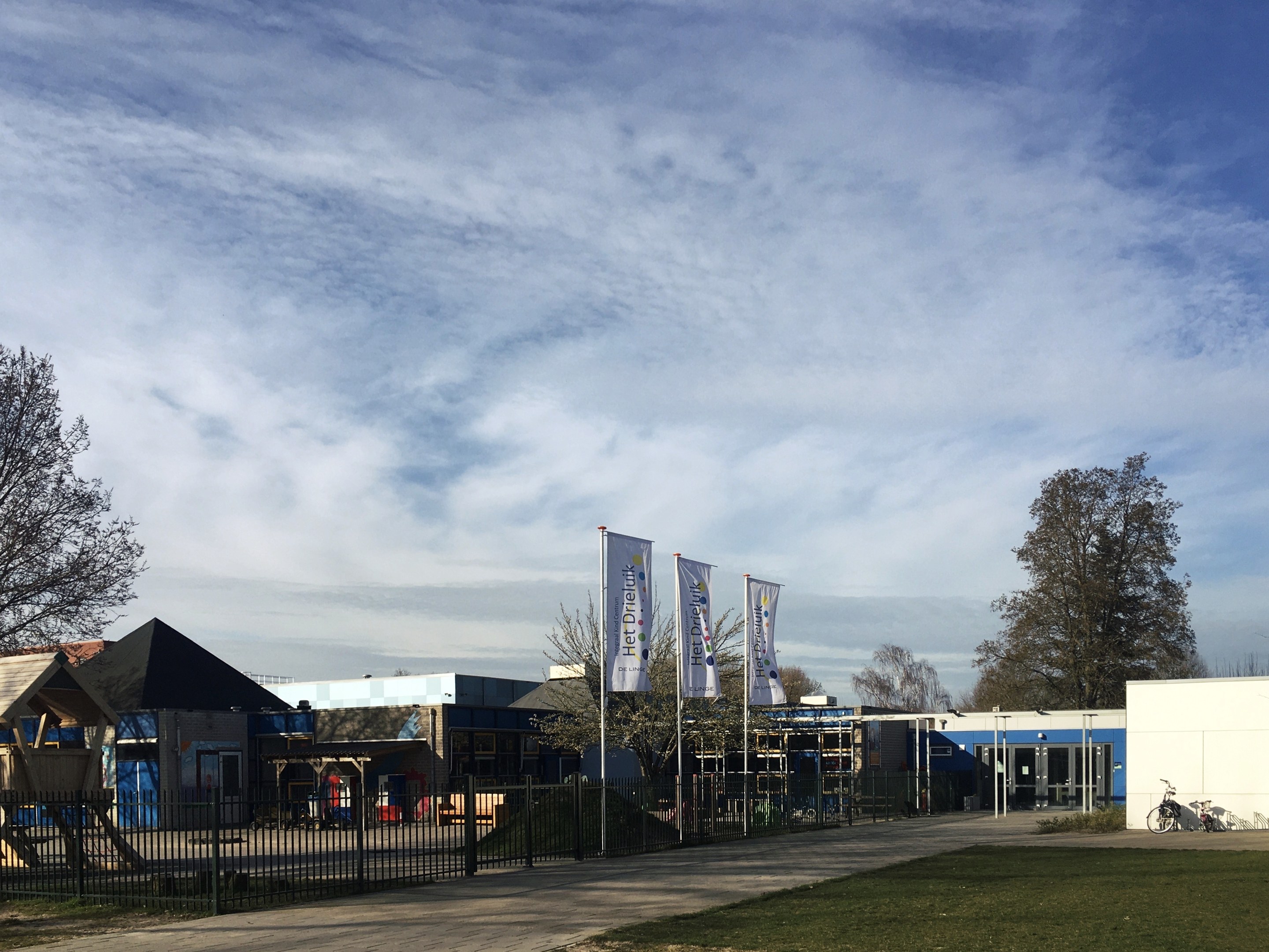 Großes Gebäude mit Metallzaun, Rasenfläche, Weg, Bäumen, Pfosten, Bannern, Fahrrad, nahegelegenen Häusern und bewölktem Himmel, als neuer Spielplatz an der Schule identifiziert.