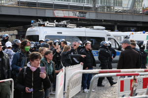 Eine Gruppe von Menschen vor Polizeifahrzeugen mit Absperrungen, einige mit Helmen und Handys, in der Nähe einer Brücke und Gebäude in Berlin während einer Protestauflösung.