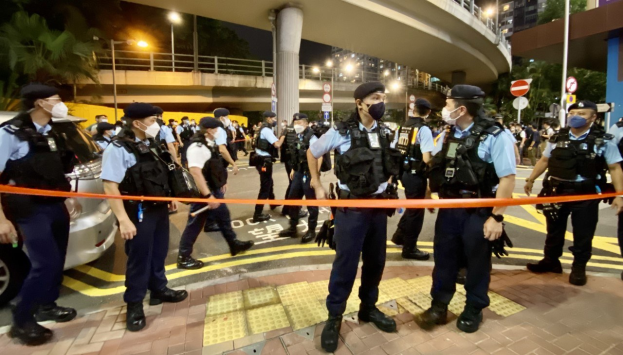 Polizeibeamte in Uniform und Masken vor einer Menge mit Fahrzeugen, Gebäuden, Bäumen und einer Brücke im Hintergrund, wahrscheinlich nach einer Demonstration in Hong Kong.