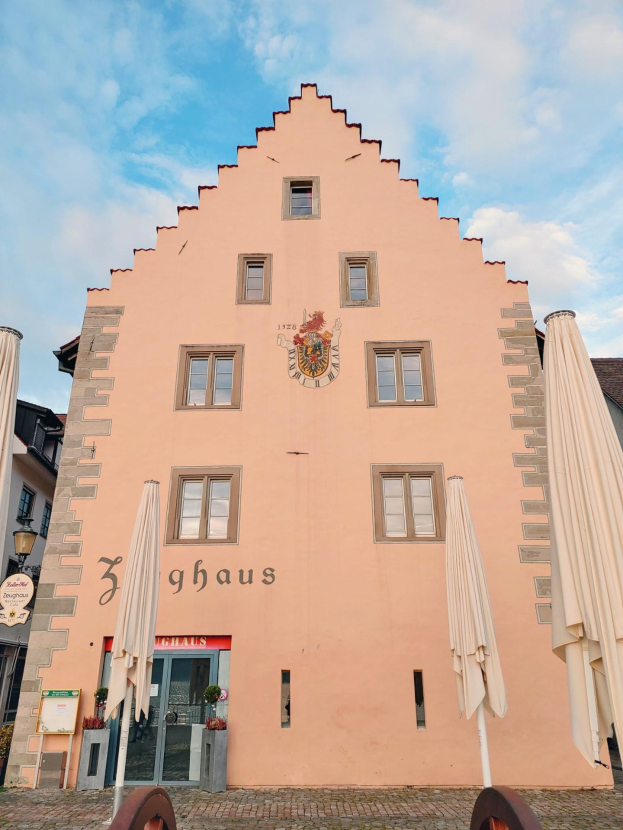 Pinkes Gebäude mit einem zentralen Glockenturm, umgeben von Fenstern, Schirmen, einem Schild, einer Straßenlaterne, Topfpflanzen und einem bewölkten Himmel, repräsentiert Sehenswürdigkeiten in München, Deutschland.