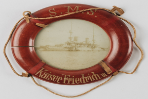 Roter Rettungsring mit 'USS Kaiser Friedrich III'-Text und Bild eines Schlachtschiffs, an einer weißen Oberfläche mit Seilen befestigt.