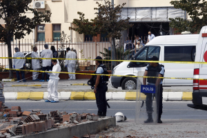 Eine Gruppe von Polizisten steht vor einem Gebäude mit Fenstern, umgeben von Fahrzeugen, einem Zaun, Bäumen und Pflanzen, mit Ziegelsteinen und einem Helm im Vordergrund, wahrscheinlich nach einer Bombenexplosion in Mexiko-Stadt.