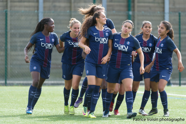 Gruppe junger Frauen beim Fußballspielen auf einem Rasenfeld mit Maschendrahtzaun und einer Wand im Hintergrund, mit dem Text "Paris Saint-Germain Frauenfußball" in der rechten unteren Ecke.