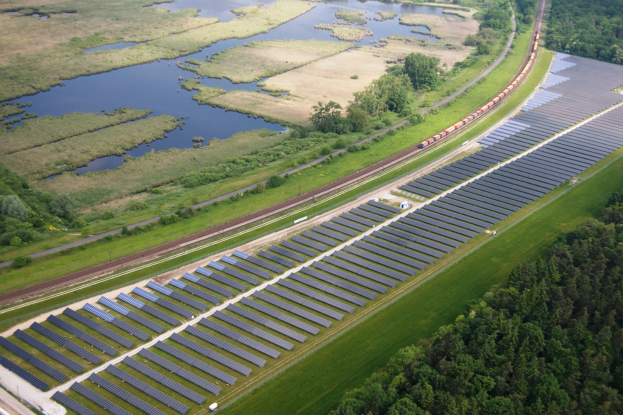 Luftaufnahme einer Solar-Farm mit Panels, umgeben von Bäumen, Gras, Wasser und einer nahen Bahnlinie.