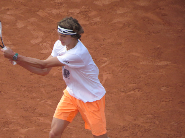 Ein Mann in einem weißen T-Shirt und orangefarbenen Shorts hält einen Tennisschläger auf einem Sandplatz und bereitet sich darauf vor, einen Ball zu schlagen.