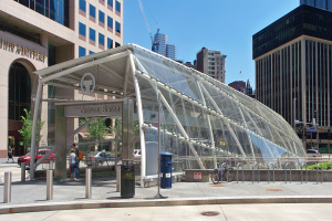 Eine belebte Stadtstraße mit hohen Wolkenkratzern, eine Bushaltestelle mit Glasdach, Fußgänger, Fahrzeuge, ein Metallzaun, ein Mülleimer, Pfosten, ein Hinweisschild, eine Ampel, Bäume, ein Kran und ein bewölkter Himmel.