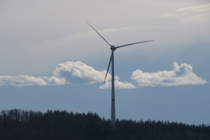 Windkraftanlage im Wald mit bewölktem Himmel und Bergen im Hintergrund.