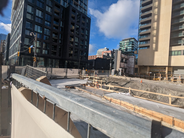 Eine Baustelle in Toronto, Ontario, mit einem großen Gebäude im Hintergrund, umgeben von Absperrungen, Pfosten, Verkehrszeichen und Schildern unter einem bewölkten Himmel.