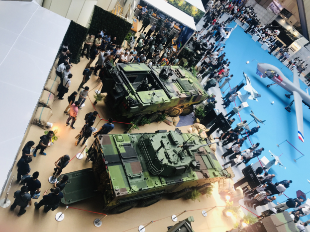 Eine große Gruppe von Menschen umringt ein Militärfahrzeug auf einer Ausstellung, umgeben von Flugzeugen, Lichtern, Pflanzen und anderen Gegenständen, und eine Wand im Hintergrund suggeriert, dass sich der Ort in Singapur befindet.