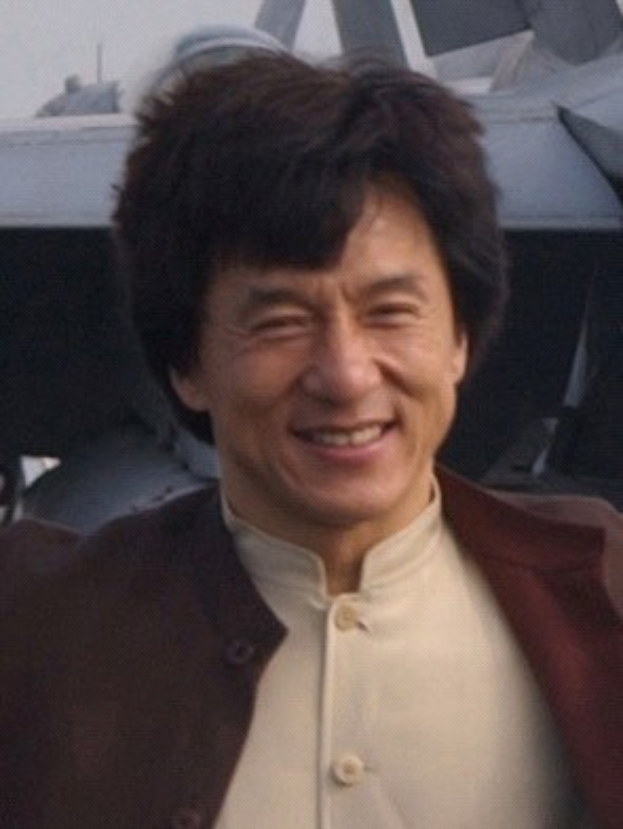 Jackie Chan steht lachend vor einem Flugzeug vor einem klaren blauen Himmel, trägt ein weißes Hemd und eine schwarze Jacke mit ordentlich gekämmtem Haar.