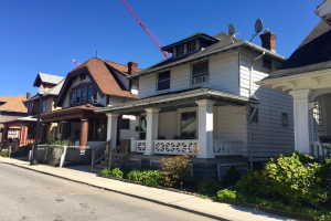 Reihe von Häusern mit Fenstern, Säulen, Geländern, Stufen und Dächern, einschließlich eines im Bau befindlichen mit einem Kran auf seinem Dach, neben einem Straßenpfahl, Pflanzen, Zaun, Weg, Bäumen und einem bewölkten Himmel.