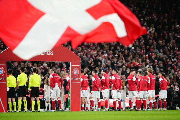 Eine Gruppe von Menschen auf einem Fußballfeld mit einer roten und weißen Flagge im Vordergrund, einem Bogen mit der Aufschrift "Bayern München vs Bayern München Betting Tips & Preview" im Hintergrund und einer großen Menschenmenge im Stadion.