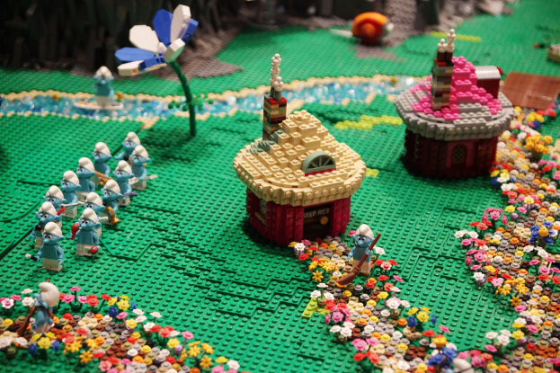 Ein LEGO-Modell einer Kleinstadt mit bunten Blumen, Häusern, Pflanzen und verschiedenen Spielzeugen, vor einem leicht unscharfen Hintergrund.