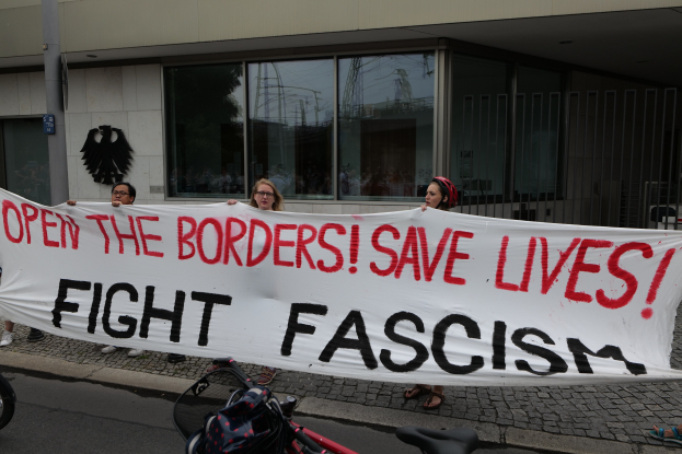 Gruppe von Menschen mit einem Transparent mit der Aufschrift "Öffnet die Grenzen, rettet Leben, bekämpft Faschismus" vor einem Gebäude mit Glasfenstern und einem Mast, mit einem Fahrrad und einer Tasche auf der Straße.