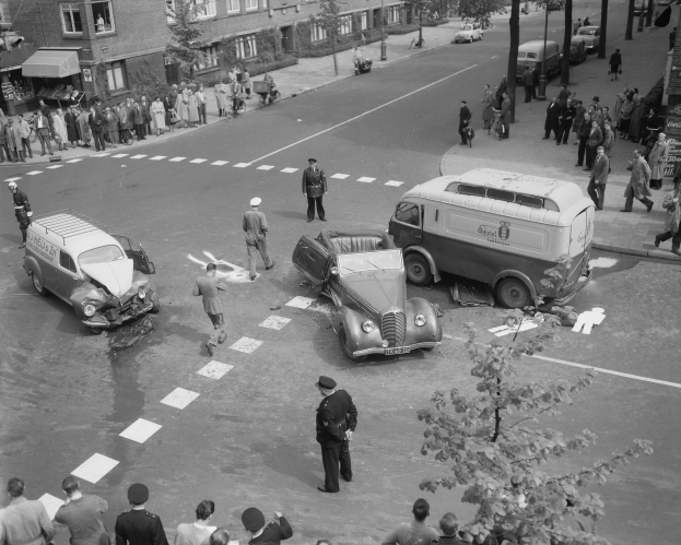 Schwarz-weiß-Szene eines Autounfalls an einer Straßenkreuzung mit mehreren Fahrzeugen, darunter ein Van, und einer Gruppe von Schaulustigen; Gebäude, Bäume, Strommasten und Schilder im Hintergrund.