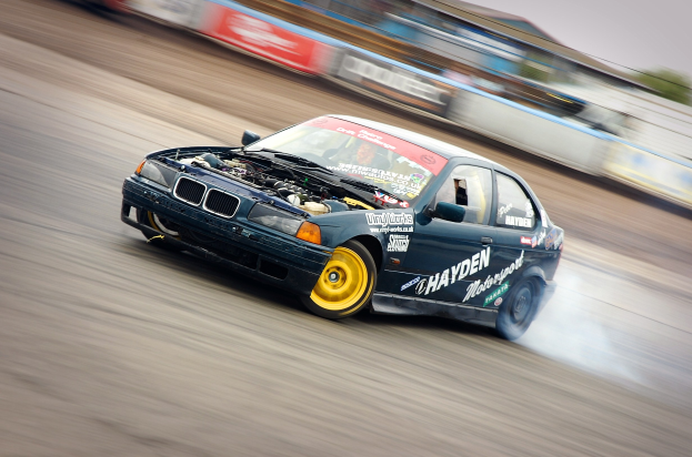 Ein Auto driftet auf einer Strecke, produziert Rauch, umgeben von Tafeln mit Text, einem Zaun, Bäumen und einem unscharfen Himmel.
