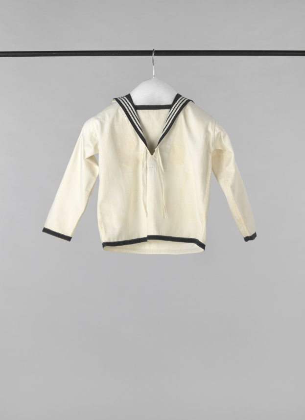 Eine weiße Matrosenjacke mit Kragen, langen Ärmeln und Knopfleiste hängt an einer Wäscheleine vor einem weißen Hintergrund.