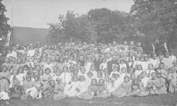Schwarz-weißes Foto von Kindern, die vor einem Haus mit Bäumen und einem klaren Himmel posieren.