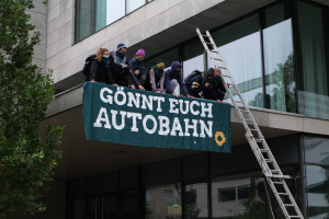 Eine Gruppe von Menschen auf dem Dach eines Gebäudes mit einer Fahne, einem Baum auf der linken Seite und Glasfenstern im Hintergrund, wahrscheinlich protestierend für die deutsche Regierung.