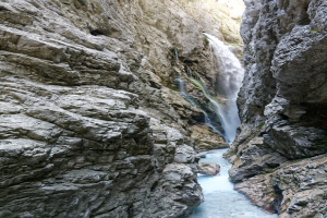 Ein kleiner Wasserfall stürzt eine steile Felsenschlucht hinab, umgeben von grünen Hügeln, mit klarem Wasser, das über raue Felsen unter strahlendem Sonnenlicht fließt.
