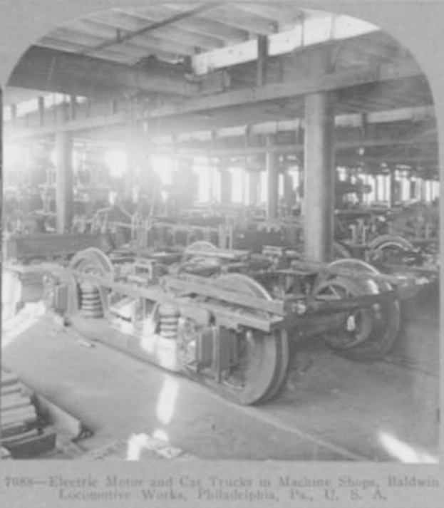 Schwarzes und weißes Foto eines elektrischen Motors und eines Lastwagens in einer industriellen Fabrikumgebung mit Maschinen, Säulen und einem Dach, mit Text unten.