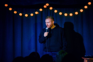 Ein Mann auf einer hell beleuchteten Bühne hält ein Mikrofon in der Hand und steht in der Nähe eines Tisches mit Gegenständen, mit einem Publikum darunter und einem Vorhang im Hintergrund, was auf eine Stand-up-Comedy-Veranstaltung hinweist.