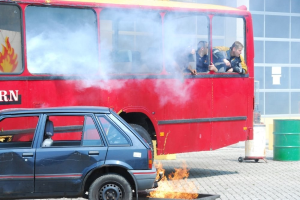 Roter Doppeldeckerbus mit Rauchentwicklung und drei sichtbaren Passagieren, neben einem Auto geparkt, vor einem Gebäude mit Glasfenstern und einem Fass auf der rechten Seite.