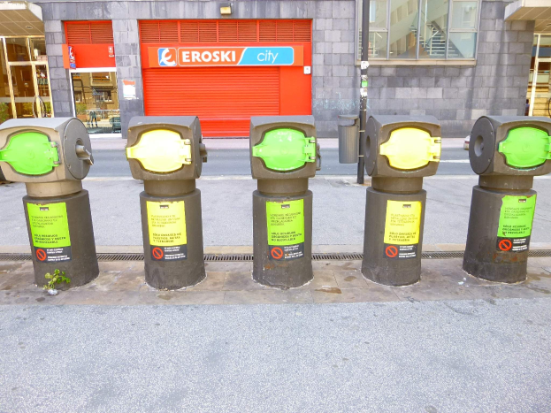 Eine Reihe von Parkautomaten entlang einer Straße mit einem Gebäude mit Glasfenstern und einem Rollladen mit Text im Hintergrund und einem nahen Müllcontainer.