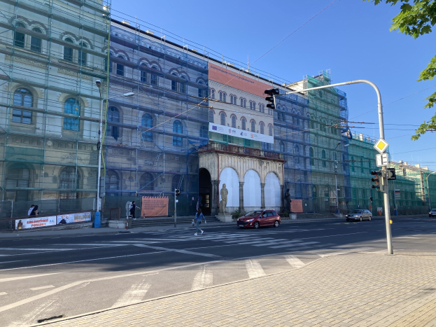 Großes Gebäude mit Gerüst während der Renovierung, umgeben von Fahrzeugen, Passanten, Ampeln, Straßenschildern, Polen, Bäumen und einem klaren blauen Himmel.