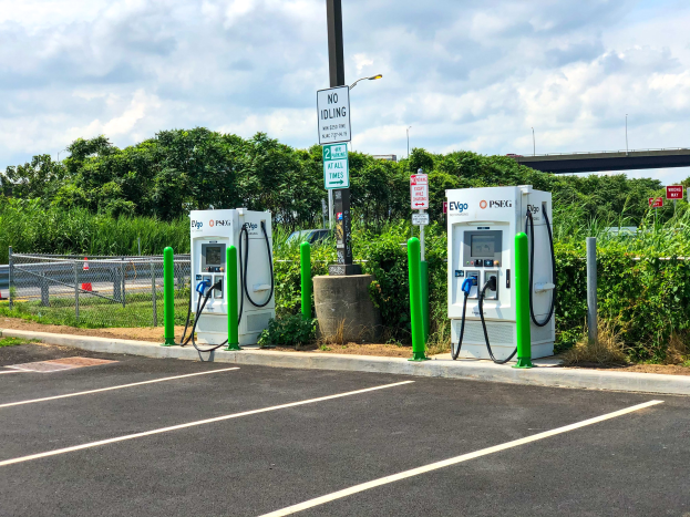 Elektrofahrzeug-Ladestation auf einem Parkplatz mit umgebender Infrastruktur, Grünfläche und einer Brücke im Hintergrund.