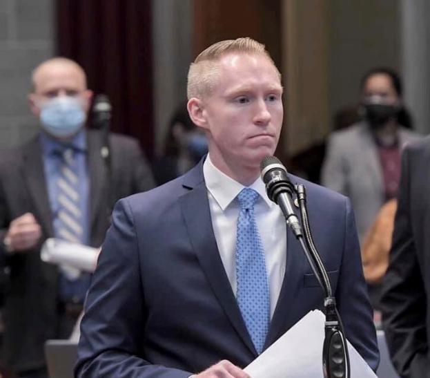 Ein Mann im Anzug spricht an einem Rednerpult mit Papieren, während er in ein Mikrofon spricht, und andere stehen in der Nähe einer Vorhangwand.