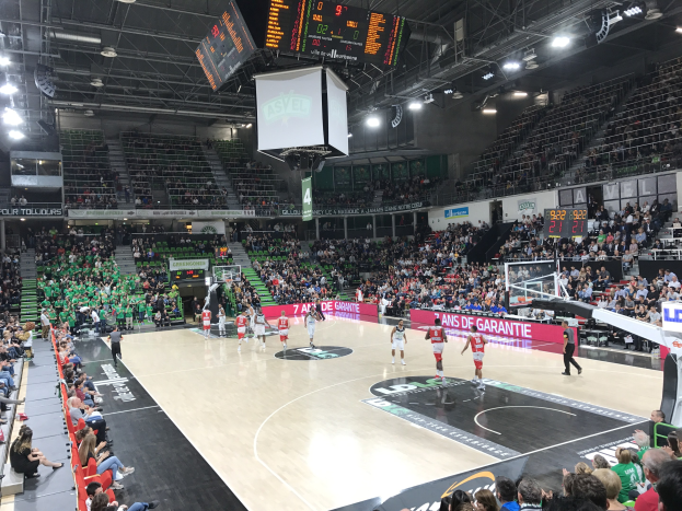 Basketballspiel in einer großen Arena mit Zuschauern, Texttafeln mit "Asvel vs Galatasaray" und Beleuchtung und Anzeigetafel.