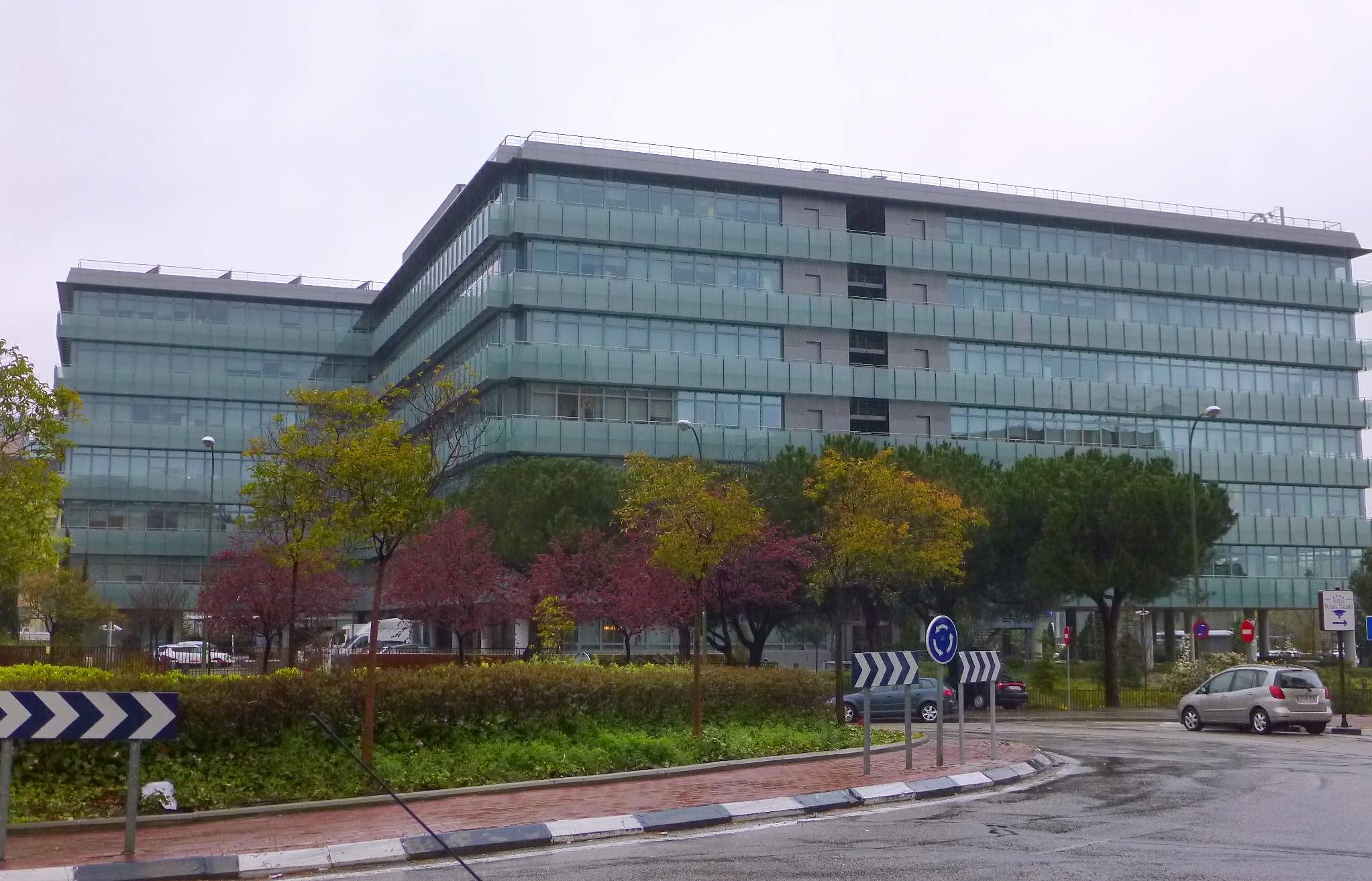 Großes modernes Gebäude mit zahlreichen Fenstern, identifiziert als Hauptsitz der Europäischen Zentralbank, umgeben von Grünflächen, Straßeninfrastruktur und Fahrzeugen unter einem bewölkten Himmel.